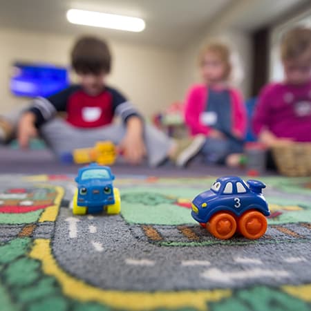 Au premier plan, deux voiturettes pour enfants posées sur un tapis représentant une route. En arrière plan, trois enfants flous.