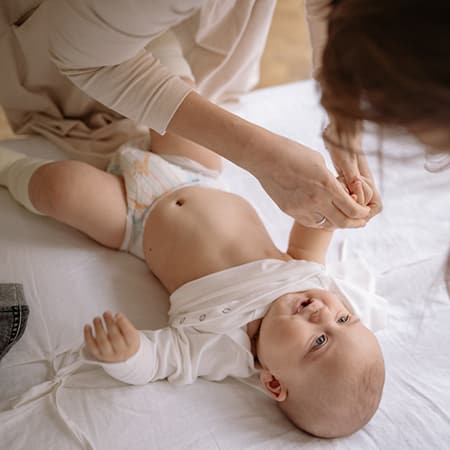 Un bébé vu par au dessus est allongé et souri à sa mère qui est entrain de l'habiller.