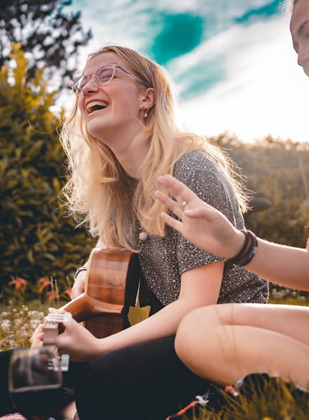 Une jeune femme rit assise dans l'herbe en jouant de la guitare.