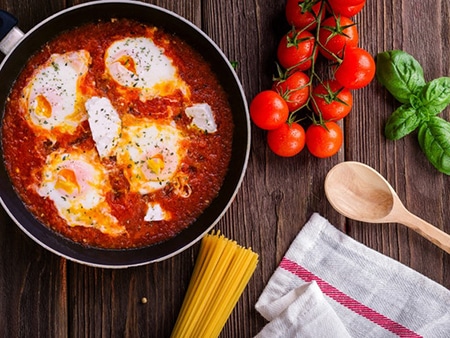 cuisine-famille-02 Dans une poêle il y a une sauce tomate avec des œufs. A coté de la poêle sont disposés des spaghettis non cuits, des tomates cerises et une cuillère en bois.