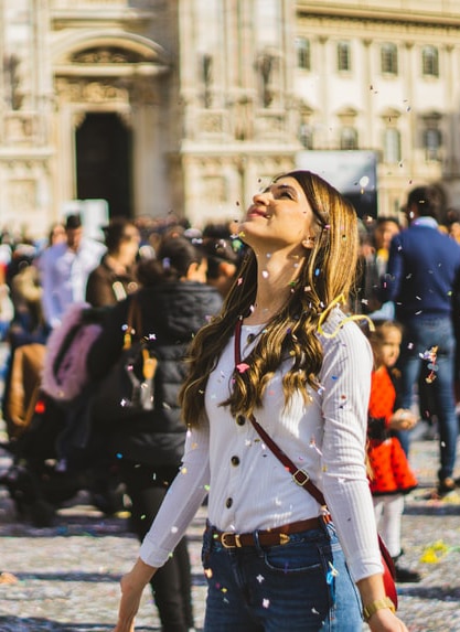 Une jeune femme, est au milieu d'une ville ou il y a du monde. Elle lève la tête en arrière avec les bras écartés. Des confettis virevoltent autour d'elle.